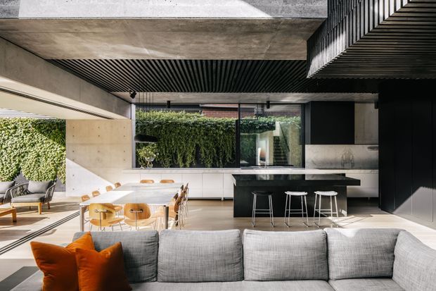 Open-plan contemporary kitchen, dining and living area with a large grey sectional sofa in the foreground. The space features a dark kitchen island with bar stools, light wood dining table and chairs, extensive glazing, a dramatic black slatted ceiling, exposed concrete surfaces, black cabinetry, and integrated vertical greenery along the rear wall. Strong natural light creates a refined indoor-outdoor atmosphere.