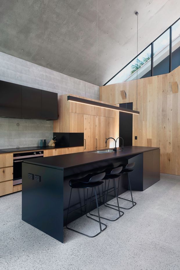 A dramatic contemporary kitchen with a large matte-black island and integrated sink, paired with black bar stools and a linear pendant light. Warm timber cabinetry and wall panelling contrast with exposed concrete walls and ceiling. High-level glazing brings in natural light, while the polished terrazzo-like concrete floor and dark cabinetry reinforce the minimalist industrial character.