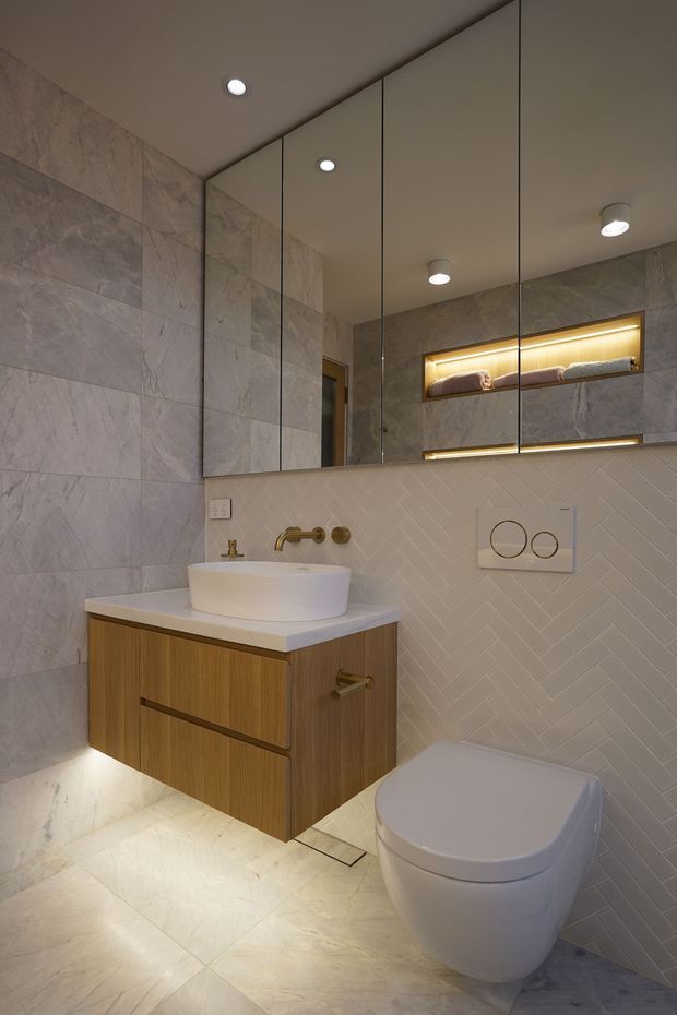A refined contemporary bathroom with a floating timber vanity, white vessel basin, brushed brass wall-mounted tapware, large mirrored cabinetry, marble-look wall and floor tiles, and a white wall-hung toilet. The vanity wall features white herringbone tiles, recessed illuminated shelving, and integrated lighting.
