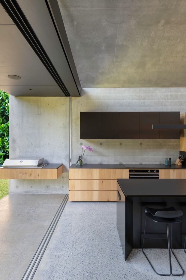 A refined modern kitchen with exposed concrete walls and ceiling, pale timber cabinetry, dark brown overhead cupboards, and a black island with integrated seating. The space opens directly to a leafy outdoor area through large glazed openings, with linear ceiling tracks, minimalist appliances, and a restrained industrial material palette.