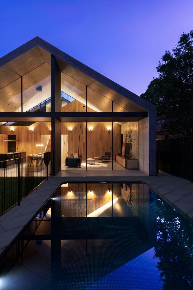 Dusk exterior view of a contemporary residence with a dramatic pitched, timber-lined roof and extensive floor-to-ceiling glazing. Warmly lit timber interiors are visible through the glass, opening onto a paved terrace beside a reflecting swimming pool. Dark metal framing, fencing and railings contrast with the warm wood surfaces and deep blue evening sky.