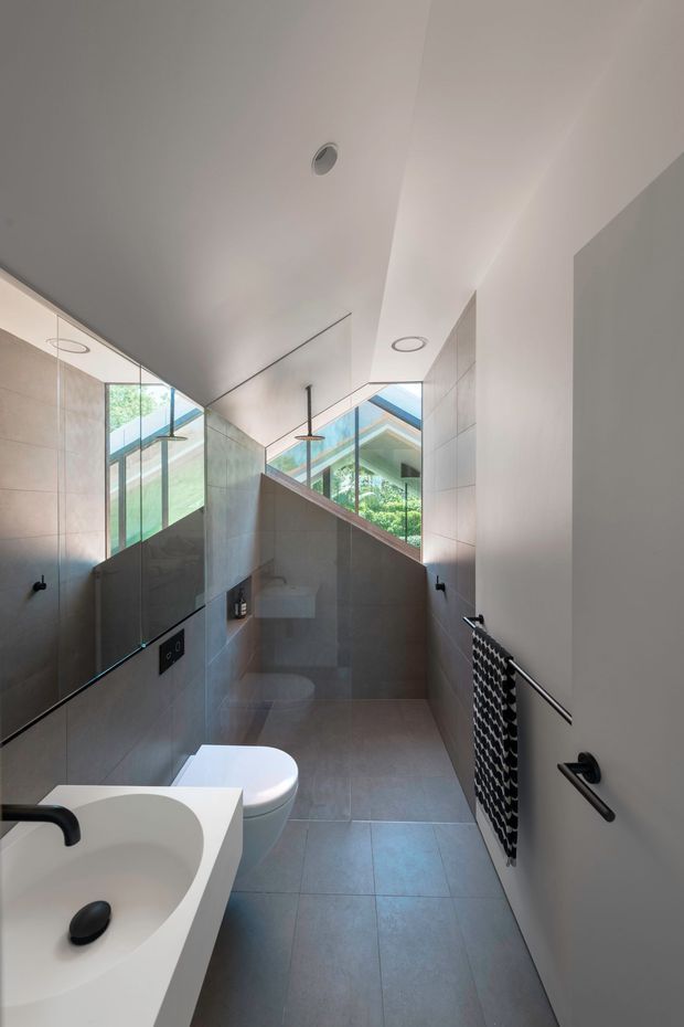 A narrow contemporary bathroom with a wall-mounted toilet, white integrated basin, large-format grey tiled walls and floor, frameless glass shower enclosure, black tapware and fittings, and a patterned black heated towel rail. A high angled window and mirrored surfaces bring in natural light and views of greenery.