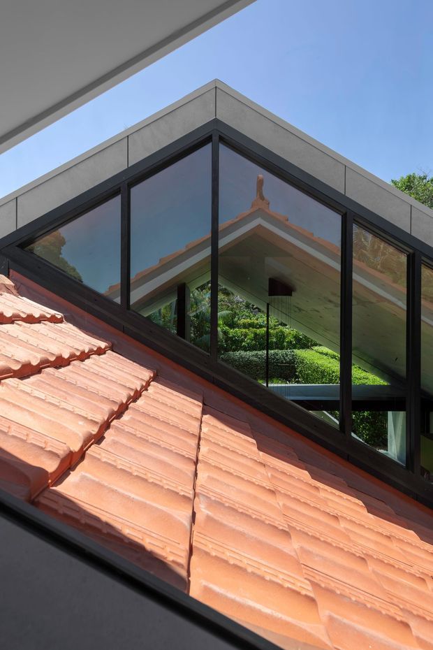 Close-up exterior architectural view of a modern glazed roof extension or upper-level corner adjoining an existing terracotta tiled pitched roof. The contemporary structure has large dark-framed glass panels and a simple grey parapet, with garden greenery and the original roofline reflected in the glazing.