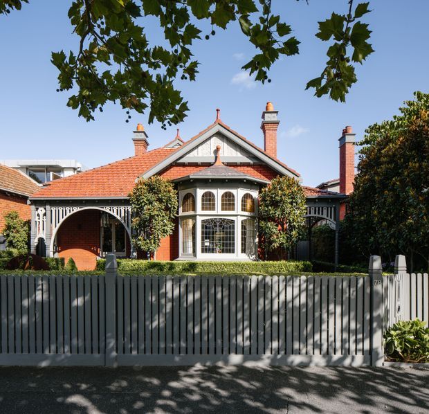 Front exterior view of a heritage-style red-brick residence with a terracotta tiled roof, multiple decorative chimneys, ornate timber verandah detailing, arched entry porch, and a prominent glazed bay window. A dark vertical-slat front fence and established hedging frame the house, with leafy branches overhead.