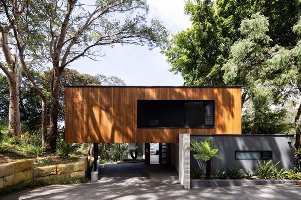 Exterior view of a contemporary elevated residential building set among mature trees and landscaped planting. The upper level is clad in warm vertical timber boards with a large dark recessed window, while the lower level combines dark painted walls, a sheltered entry or carport-like opening, and a pale textured masonry feature wall. Sandstone retaining blocks and a paved forecourt complete the scene.