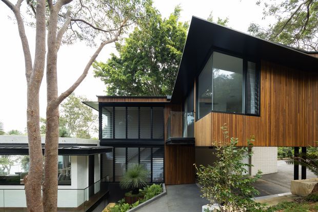 Exterior view of a contemporary multi-level residence set among mature trees. The composition features angular projecting volumes, dark metal roof planes, extensive glazing, vertical timber cladding, white masonry elements, and a covered lower-level connection. Landscaped planting and a paved approach surround the building.