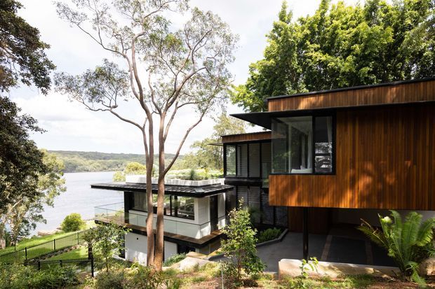 Exterior view of a contemporary multi-level home on a sloping, wooded waterfront site. The building features projecting flat roof planes, extensive floor-to-ceiling glazing, dark metal elements, and warm vertical timber cladding, with landscaped terraces stepping toward the water.