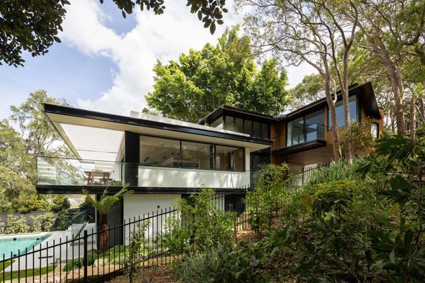 A contemporary multi-level house set within dense landscaped vegetation. The building has strong horizontal and cantilevered forms, extensive glazing, dark metal framing, pale rendered walls, and an upper-level terrace or balcony. A swimming pool and landscaped garden are visible at the lower left, while mature trees surround the property.
