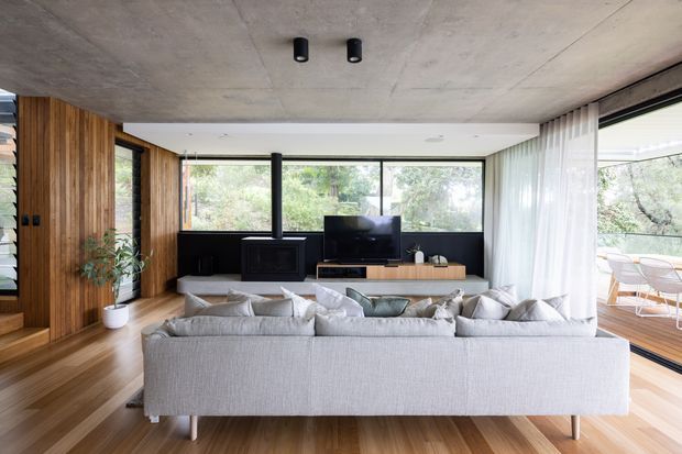 A bright, contemporary living room with a large light-grey sectional sofa facing a low timber media console and television. The room features warm timber wall panelling and flooring, expansive black-framed glazing with sheer curtains, a black fireplace or wood-burning stove, and a raw concrete ceiling. An outdoor dining area is visible through the glazing.