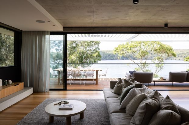 A contemporary living room opening onto a timber outdoor deck with expansive floor-to-ceiling glazing and views across a calm body of water. The room features a large grey sectional sofa, round white coffee table, low timber media console, neutral rug, sheer curtains, dark window frames, and a concrete ceiling. A dining area and outdoor seating are visible beyond the glazing.
