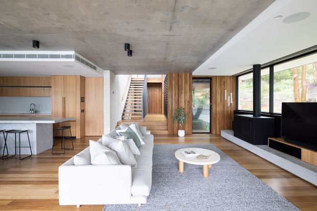 Open-plan contemporary living area with an integrated kitchen, timber cabinetry and wall panelling, polished concrete ceiling, timber flooring, and a staircase leading upward at the centre. A large white sectional sofa faces a black fireplace and media unit, with expansive black-framed glazing to the right.