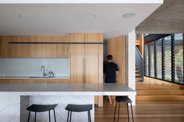 Contemporary open-plan kitchen and circulation area with extensive warm timber cabinetry, a pale stone island with waterfall edge, and black bar stools. A white tiled or brick-lined stair zone and large dark-framed louvred glazing are visible to the right, with timber flooring and stairs continuing through the space.