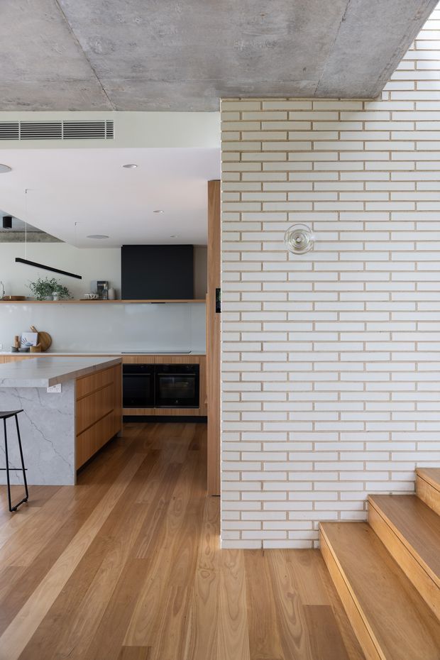 A contemporary open-plan kitchen viewed from the adjacent stair area. The space features a white brick partition wall, exposed concrete ceiling, warm timber flooring and stair treads, timber kitchen cabinetry, and a pale stone island benchtop. Black appliances and fixtures provide contrast, while a partial-height kitchen wall opens the room visually to the living area.