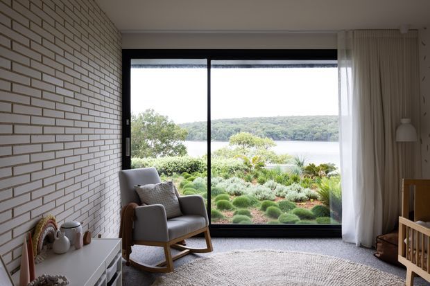 A calm, light-filled living room with a large black-framed floor-to-ceiling window overlooking a landscaped garden and lake. The room features a white painted brick feature wall, pale neutral furnishings, a textured round rug, sheer and full-height curtains, a grey rocking armchair, and a low white console. A timber cot is partially visible at right, suggesting a nursery or family living setting.