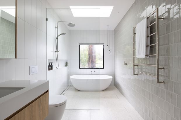 Bright contemporary bathroom with a freestanding white bathtub positioned beneath a rear window, an open shower with dual showerheads, wall-mounted basin vanity, partially visible toilet, heated towel rail, and pale grey glossy rectangular wall tiles. A large skylight provides additional natural light.
