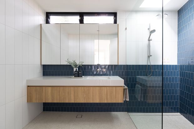 Bright contemporary bathroom featuring a wall-mounted timber vanity with a pale benchtop, mirrored cabinetry, white wall tiles, and a walk-in shower enclosed by frameless glass. Deep blue tiled walls create a strong feature backdrop, while a skylight and high windows provide abundant natural light.