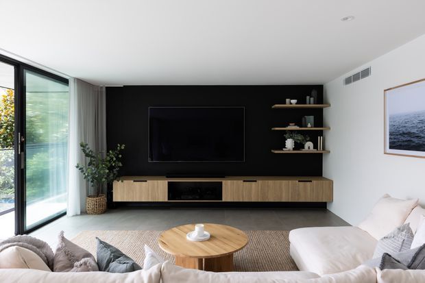 Bright contemporary living room viewed from behind a large sectional sofa. The room features full-height glazed doors or windows at left, a floating timber entertainment console with black inset cabinetry, a matte black TV feature wall, open timber shelving, neutral furnishings, a round timber coffee table, and a framed ocean photograph.