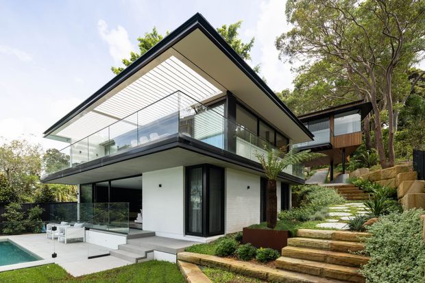 Contemporary multi-level residential home set into a landscaped, sloping site. The building features crisp white rendered walls, dark framed glazing, extensive glass balustrades, broad cantilevered flat roof planes, and a partially covered upper terrace with a linear pergola. Stone steps and planted terraces lead up to a timber-clad wing beneath mature trees, while a small pool is visible at the lower level.