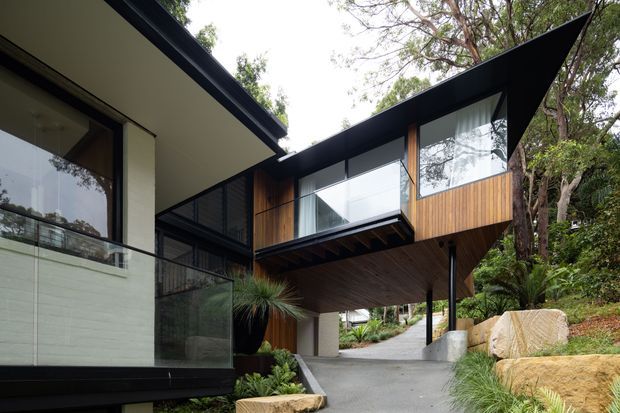 Exterior view of a contemporary split-level residence set among established vegetation on a sloping site. The building features dramatic angular roof planes, extensive glazing, dark metal framing, warm timber-clad projecting volumes, exposed structural elements, and a covered driveway or entry passing beneath the upper storey. Natural stone retaining elements and landscaped planting frame the approach.