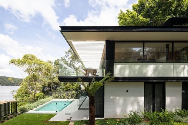 Modern two-storey house on a landscaped waterfront or lakeside site, featuring a strong flat roof, deep eaves, black-framed glazing, white wall surfaces, and a projecting upper-level terrace with clear glass balustrades. A swimming pool and outdoor terrace sit beside the house, with water and wooded hills visible beyond.