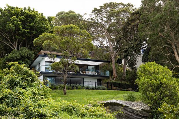 A contemporary multi-level home nestled among mature trees and landscaped gardens. The building features dark metal roofing, white and charcoal facade elements, extensive glazing, broad projecting rooflines, and sheltered outdoor terraces. A gently sloping lawn, dense planting, and large natural rocks surround the house.