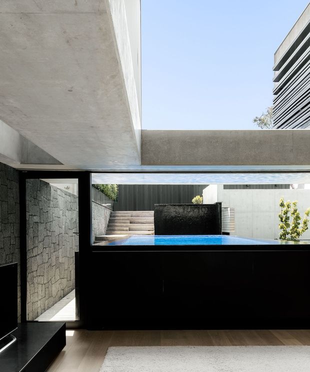 A contemporary interior opening onto a compact courtyard pool. The space features a dramatic exposed concrete overhead slab and beam, black-framed glazing, dark built-in cabinetry or wall panelling, a textured dark stone wall, and warm timber flooring. Through the glazing, blue pool water, concrete steps, rendered walls, and planted greenery are visible. The composition is minimalist and strongly connected to the outdoors, with high contrast between charcoal architectural elements, pale concrete, timber, and blue water.