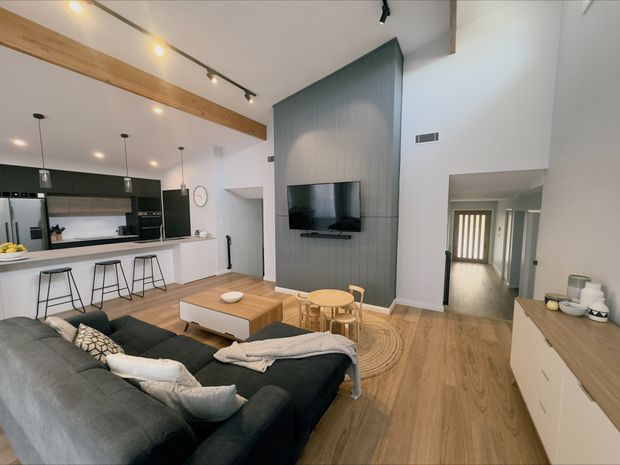 Open-plan contemporary living area with a kitchen and breakfast island at left, a large dark sectional sofa in the foreground, a timber coffee table, children's or occasional stools, and a wall-mounted television set into a tall grey vertical-panel feature wall. Warm timber flooring and exposed timber ceiling beams contrast with the white walls and dark kitchen cabinetry. A hallway leads toward the rear of the home.