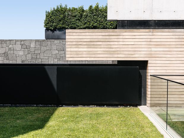 Exterior detail of a contemporary residence featuring horizontal light timber cladding, a dark metal gate and wall elements, a textured grey stone boundary wall, a crisp white rendered upper volume, and frameless glass balustrading beside a neatly landscaped lawn.