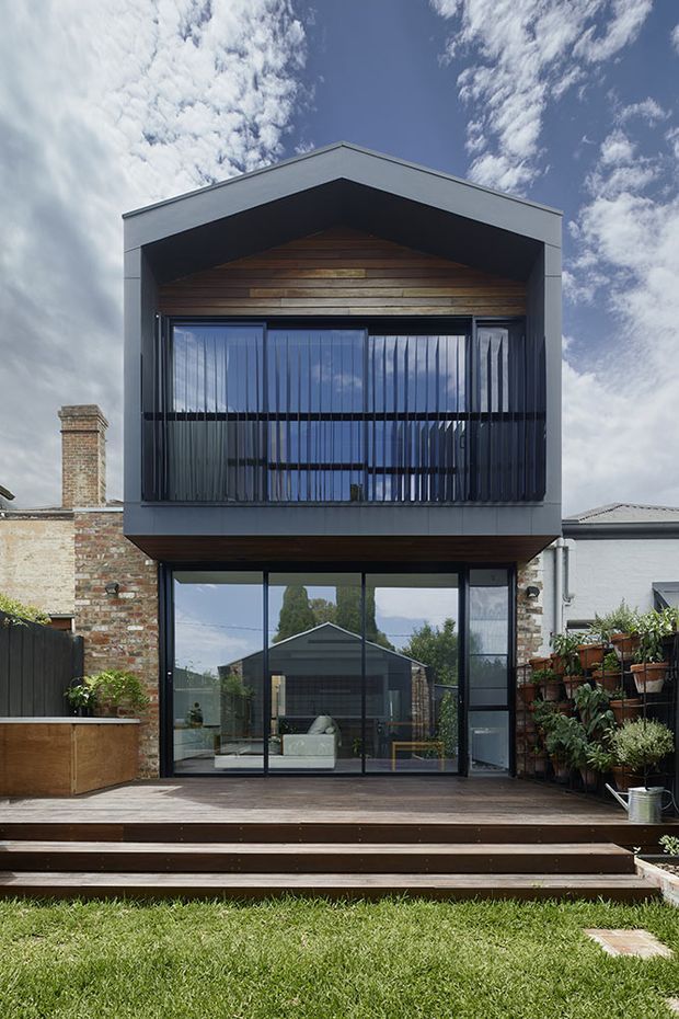Front-facing exterior view of a narrow contemporary two-storey residential extension or townhouse. The facade has a strongly geometric gable-like projecting form, dark grey metal cladding, extensive black-framed glazing, and a timber-lined upper balcony recessed behind vertical metal balustrades. A timber deck and steps lead to the glazed ground-floor living area, with brickwork and neighbouring residential structures visible at the sides. The image is photographed in daylight from the garden.