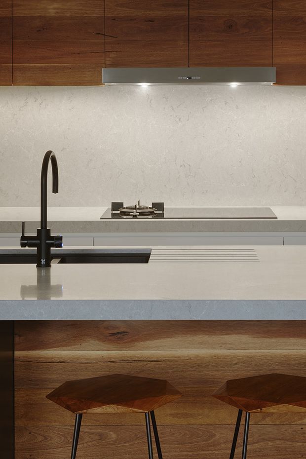 Close-up architectural photograph of a refined contemporary kitchen. Dark natural timber cabinetry and matching timber wall panelling frame a pale stone or engineered-stone benchtop and backsplash. A slim stainless-steel rangehood, black gooseneck tap, integrated cooktop and linear draining grooves create a clean, minimalist composition. Two timber bar stools with black metal legs are visible below the island.