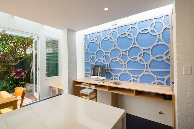 Bright open-plan interior featuring a compact dining and kitchen area, with a built-in timber study or media desk along the wall. The main feature is a large blue decorative circular-pattern screen or panel set against a white brick wall. White cabinetry and benchtops, timber joinery, recessed lighting, and glazing opening to a leafy courtyard create an airy contemporary setting.