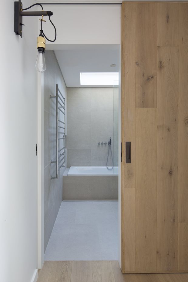 A compact contemporary bathroom viewed from the hallway through a partially open sliding timber door. The bathroom features pale grey large-format tiled walls and floor, a built-in bathtub with wall-mounted fixtures and handheld shower, a stainless heated towel rail, and a rectangular skylight. A black-and-brass exposed-bulb wall light is mounted outside the bathroom, while warm natural timber flooring and the large timber door add contrast.