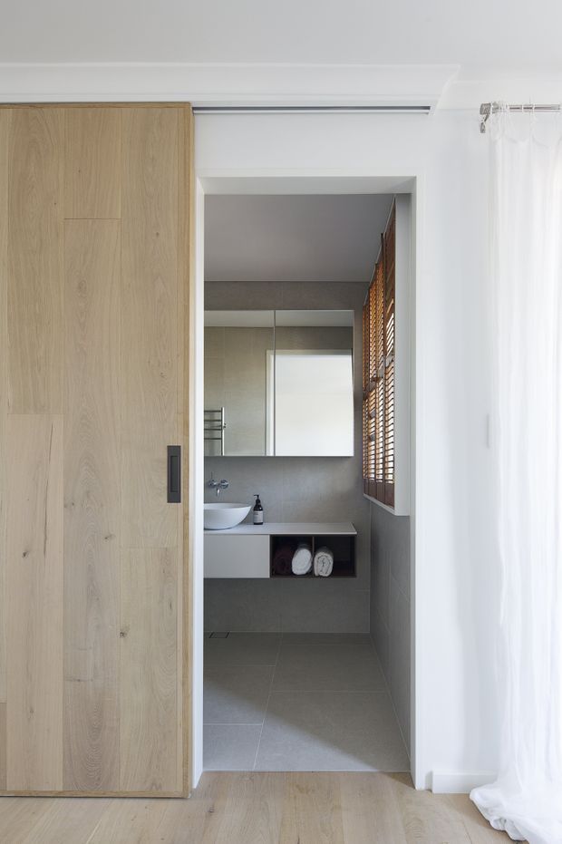 A bright, minimalist bathroom viewed from an adjacent bedroom or dressing area. The compact bathroom features large grey floor and wall tiles, a floating white vanity with a round vessel basin, wall-mounted tapware, a broad mirrored cabinet, and open towel storage. A light natural timber sliding door partially conceals the bathroom, while timber Venetian blinds and sheer white curtains provide warmth and filtered daylight. The surrounding floor is pale timber, and the space has clean contemporary detailing.