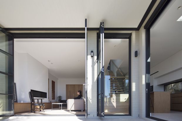 Bright contemporary interior view through large black-framed glazed doors, showing a living room on the left, a central stairway with open timber treads, and part of a kitchen on the right. White walls and ceilings, pale flooring, dark window frames, and extensive glazing create an airy, connected space.