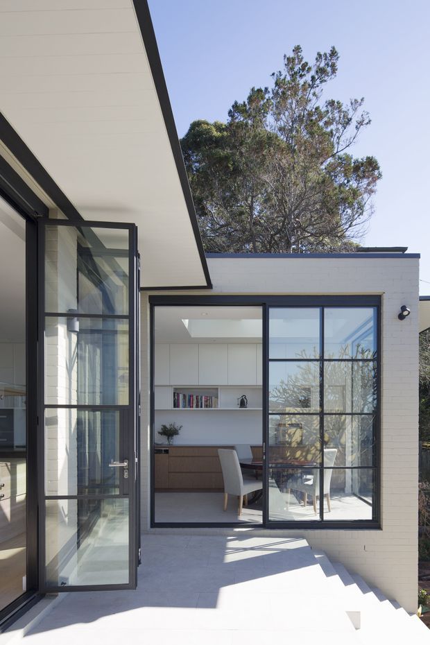Exterior view of a contemporary residential extension with a bright white-painted brick facade, broad flat roof overhang, black-framed steel-look glazing, and an open glazed door leading onto a pale paved terrace. Through the windows, a dining area and built-in timber cabinetry are visible. Mature trees surround the house, and strong sunlight creates sharp shadows across the terrace.