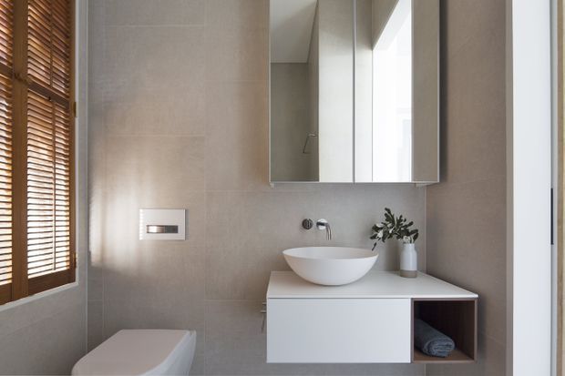 Contemporary compact bathroom featuring warm grey full-height wall tiling, a white vessel basin on a minimalist floating vanity, mirrored wall cabinetry, wall-mounted tapware, and an integrated toilet partially visible at left. Timber Venetian blinds provide warmth beside the vanity, while a small potted plant adds a natural accent.