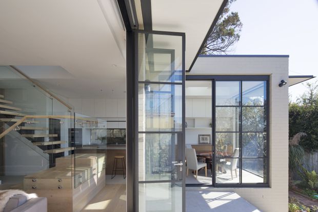 A bright contemporary residential interior viewed through a glazed connection between living and work areas. The image features extensive black-framed glazing and a glazed pivot door, pale timber built-in cabinetry, a glass balustrade with floating timber stair treads, and a compact home office beyond. Strong daylight creates an airy, open feel, with a small landscaped exterior visible through the windows.