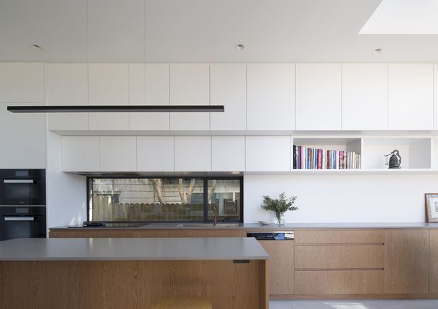 A bright, contemporary minimalist kitchen with full-height white cabinetry, integrated black appliances, a long linear black pendant light, and a timber island with a pale benchtop. Matching timber cabinetry continues along the back wall beneath open white shelving displaying books and decor. A wide window above the cooktop brings in natural light, while a skylight adds overhead illumination.