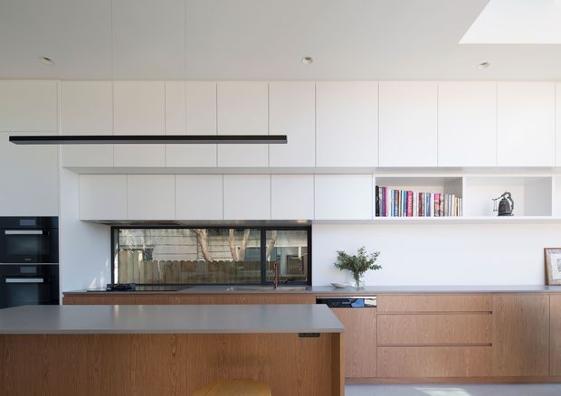 A bright, contemporary kitchen with a large island, white full-height cabinetry, integrated black appliances, a long horizontal window, and warm timber base cabinetry. Open shelving displays books and minimal decor, while a linear black pendant light and concealed ceiling lighting provide a clean architectural finish.