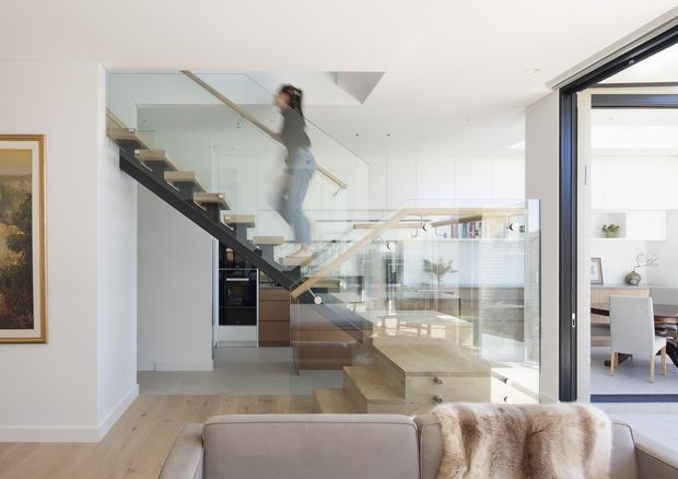 Bright contemporary open-plan interior viewed from a living area, featuring a floating timber staircase with a dark structural stringer, clear glass balustrades, and timber handrails. Pale timber flooring continues through the space, with white walls, extensive glazing, and a glimpse of a dining area beyond. A sofa with a fur throw occupies the foreground, while built-in joinery and a kitchen-like area are visible behind the staircase.
