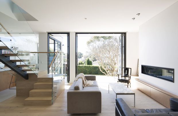 Bright contemporary living room with a minimalist built-in television wall and linear fireplace, opening to a landscaped garden through large glazed doors. A timber staircase with clear glass balustrade and open shelving occupies the left side, while neutral sofas and a low coffee table create a calm seating area.