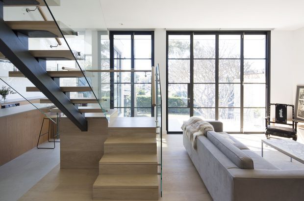 Bright contemporary open-plan interior featuring a sculptural timber stair with dark steel stringers and glass balustrades, opening into a living room with a large light-grey sofa. Full-height black-framed glazed doors and windows bring in abundant daylight, while pale timber flooring and joinery create a clean, warm setting.