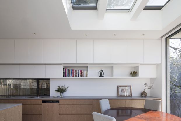 Bright contemporary open-plan kitchen and dining area with extensive white overhead cabinetry, warm timber base cabinetry, a dark stone or glass-like benchtop, integrated shelving, and a polished dining table. Large skylights and full-height glazing provide abundant natural light, while a neutral palette and clean-lined joinery create a refined, uncluttered interior.