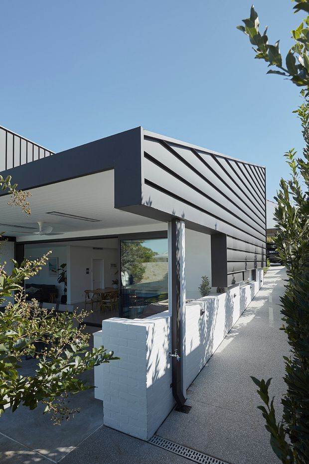 Exterior view of a contemporary single-storey residence with a bold projecting flat roof and dark horizontal metal cladding. The home features an open covered outdoor living area, large glazing, white painted brick walls, slender dark structural columns, and a paved side walkway with linear drainage. Mature planting frames the building.
