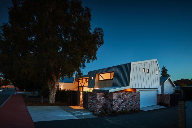 Twilight exterior view of a contemporary multi-level residence with a sharply angled corrugated metal upper volume, exposed timber framing, red-and-white brick lower walls, and an integrated white garage door. A large mature tree frames the driveway and pedestrian path, while warm interior lighting glows through the windows.