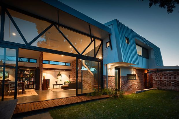 Twilight exterior view of a contemporary multi-level home featuring extensive floor-to-ceiling glazing, exposed black structural framing, red brick walls, and a distinctive corrugated blue-grey metal upper facade. A timber deck opens to a warmly lit interior with visible living and dining areas, bookshelves, and planted features. The home has a strong indoor-outdoor connection and a landscaped lawn in the foreground.