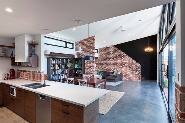Open-plan loft-style kitchen and living area with a large white island, dark timber cabinetry, exposed red brick walls, polished concrete flooring, extensive floor-to-ceiling glazing, a built-in bookshelf, and a dramatic black feature wall. Pendant lights hang above the island, while a grey sofa and rug define the living zone.