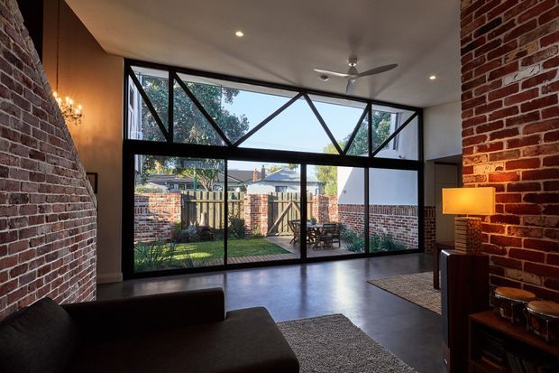 Industrial-style living room with exposed red-brick walls, polished dark flooring, and a large wall of black-framed glazing opening visually onto a small brick-enclosed courtyard. The glazing includes prominent diagonal steel bracing and high-level clerestory-like panels. A dark sofa, rug, floor lamp, chandelier, ceiling fan, and compact timber media/storage unit furnish the room.
