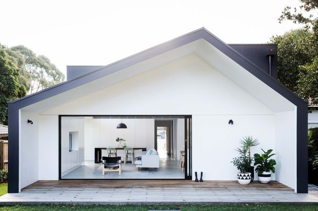 A bright, single-storey gabled house extension or pavilion with a crisp white facade, dark grey roof and trim, and a large black-framed glazed opening. A timber deck runs across the front, while the interior reveals an open-plan living and dining area with contemporary furnishings. Potted plants and simple exterior wall lights flank the glazing.