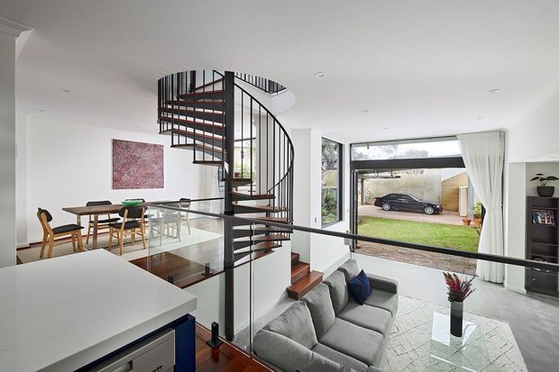 Bright open-plan contemporary living space with a sculptural black metal spiral staircase, timber stair treads, and glass balustrade. A grey sectional sofa sits beside a pale stone or concrete-look coffee table, while a dining area and kitchen island occupy the foreground and left side. Large windows and glazed doors open the room toward a small lawn and driveway, creating an airy connection to the outdoors.